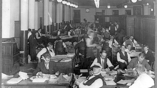 A black and white image of the City room of the New York World Magazine, circa 1900. Men in old fashioned suits sit at desks covered in paper in a long room with columns along one wall.